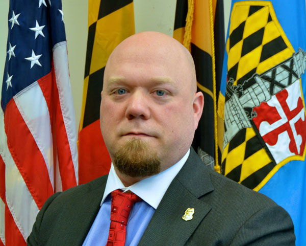 Steven O'Dell standing in front of the American, Maryland, and Baltimore flags. 