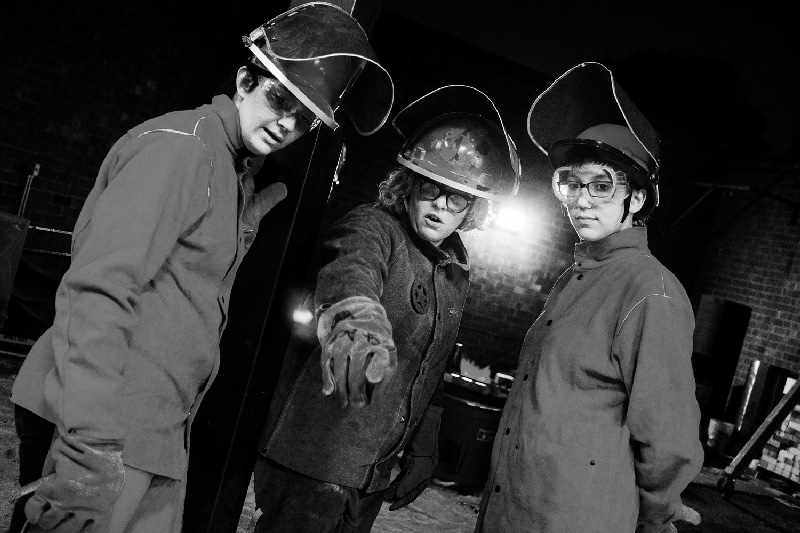 Stacey Holloway in coveralls and a welding helmet and gloves, teaching two similarly attired students.