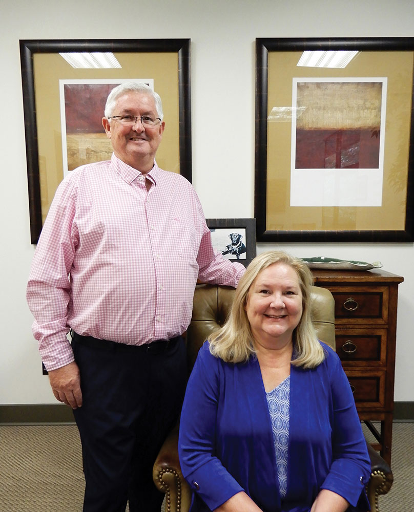 Phyllis Brasfield sits in an office chair, David Brasfield stands behind her resting his hand on the top of the chair. 