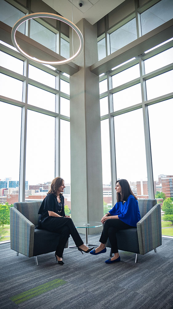 Shaia and Khan sitting in chairs facing each other; behind them is a wall made of glass and campus buildings. 