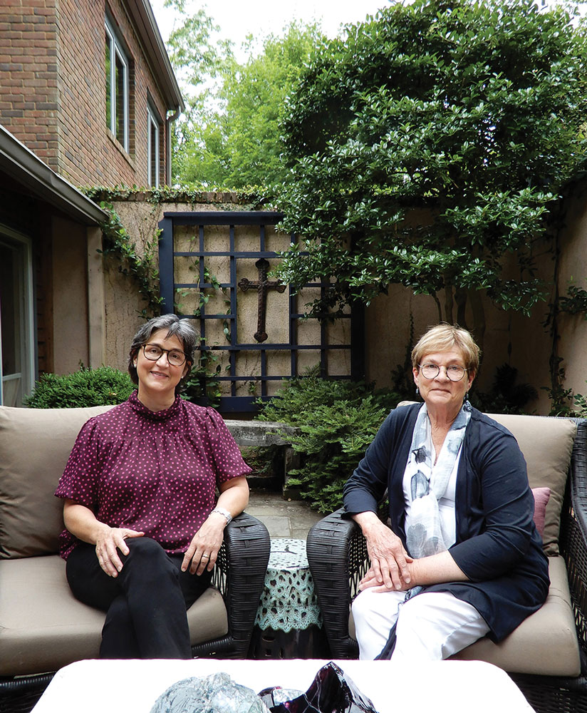 Alison and Karen Chapman sitting in wicker chairs on a stone patio, plants and a cross behind them.