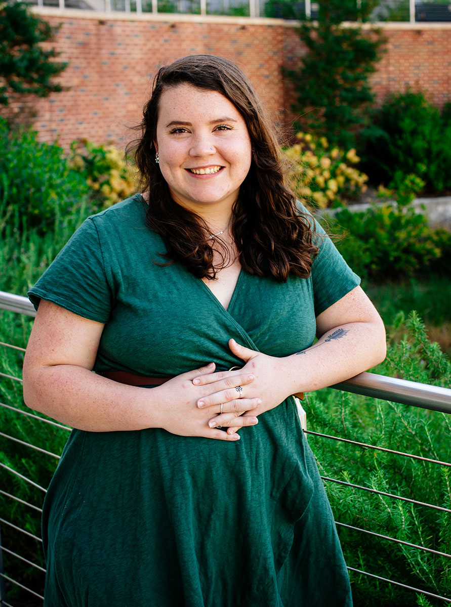  Lexie Woolums, smiling and wearing a green dress, leans on a railing in front of a garden and a brick building. 