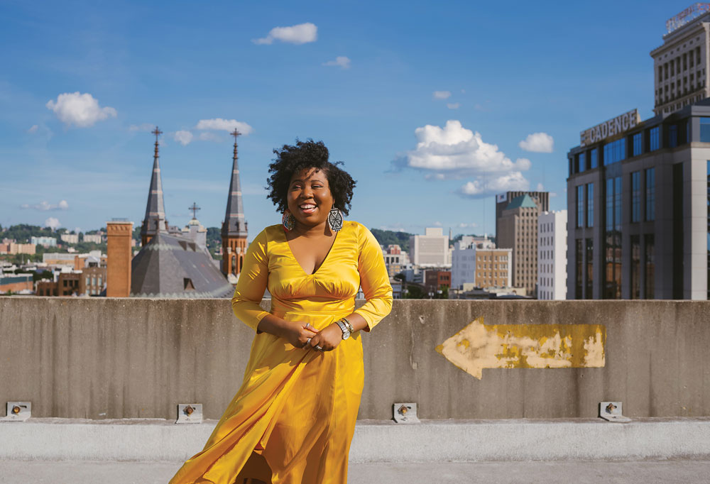 A wider shot of Ashley on the roof, wearing a yellow dress under a bright blue sky with a few white clouds. 
