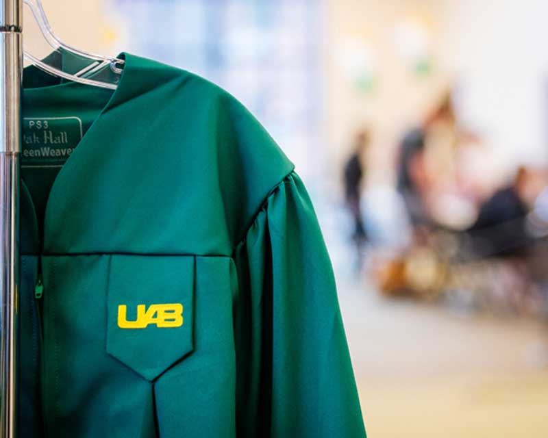 A UAB graduation gown on a hanger with students in the background.