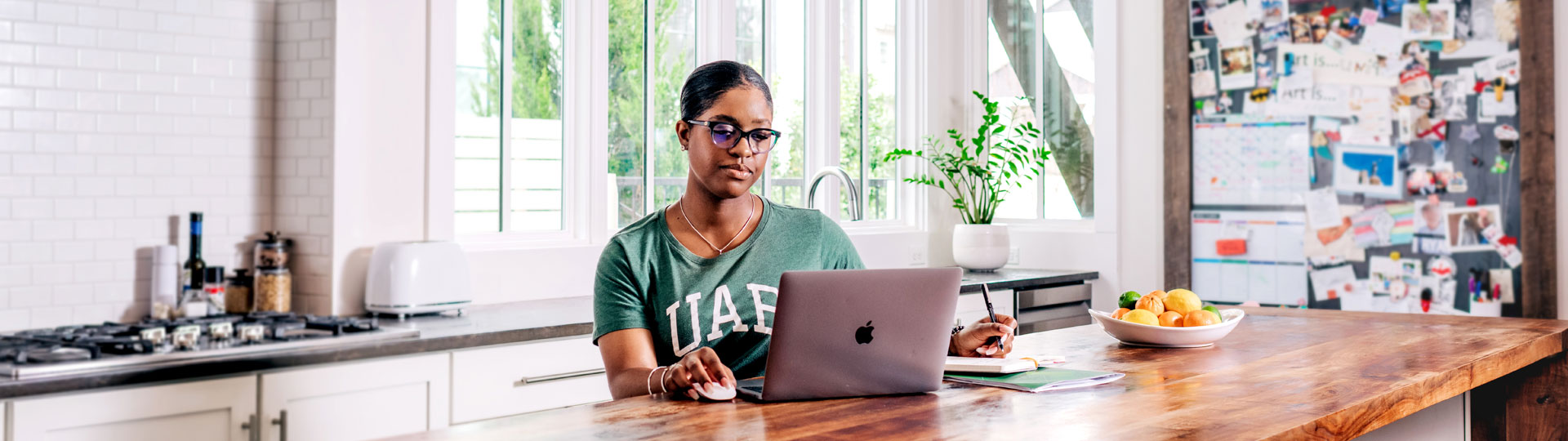 Black female at her laptop in a window-filled kitchen, wearing a UAB tshirt.