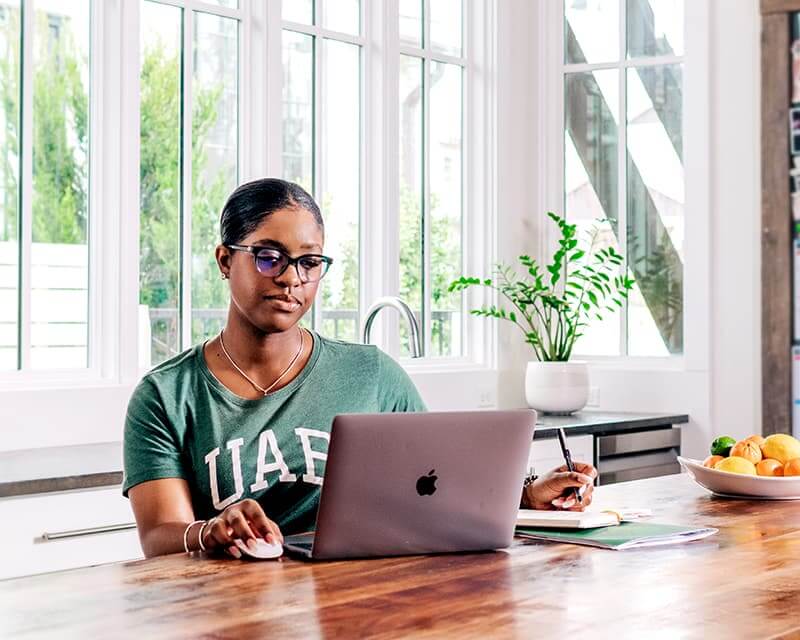 Black female at her laptop in a window-filled kitchen, wearing a UAB tshirt.