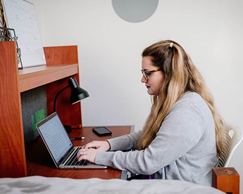 Caucasian female with long blond hair and glasses in a gray sweater, working at a laptop.