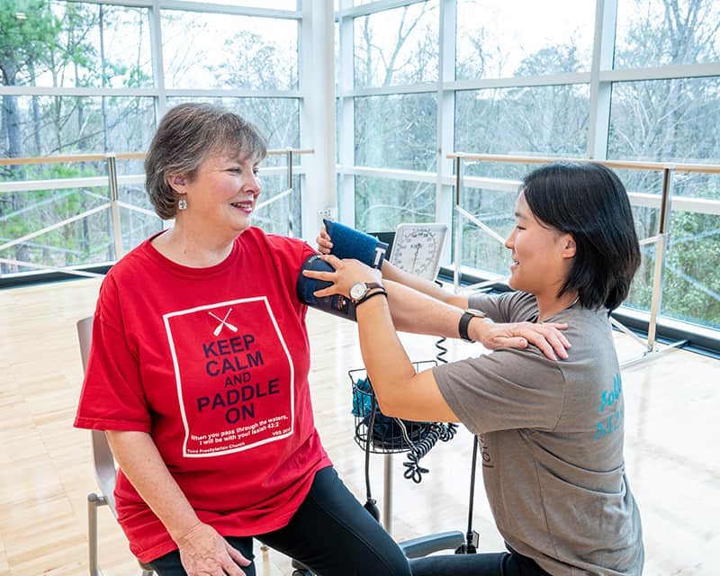 An Asian woman checks the blood pressure of an older white woman wearing a red t-shirt that reads "keep calm and paddle on." 