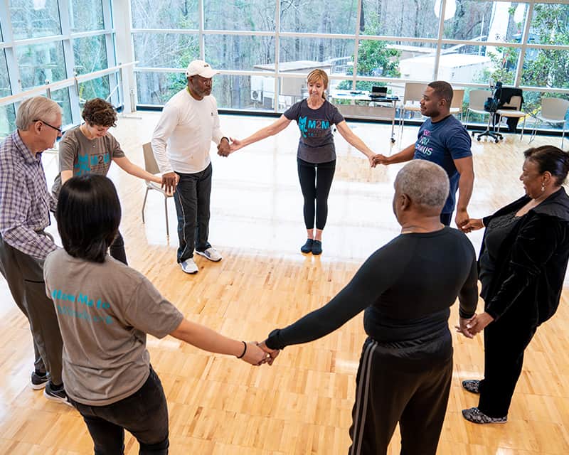 Instructor and group exercise class participants hold hands in a circle in windowed classroom. 