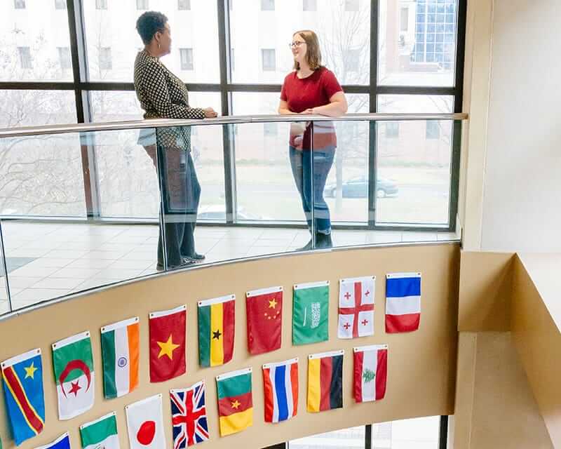 Two women talking on a balcony with glass wall,  international flags displayed on the wall below. 