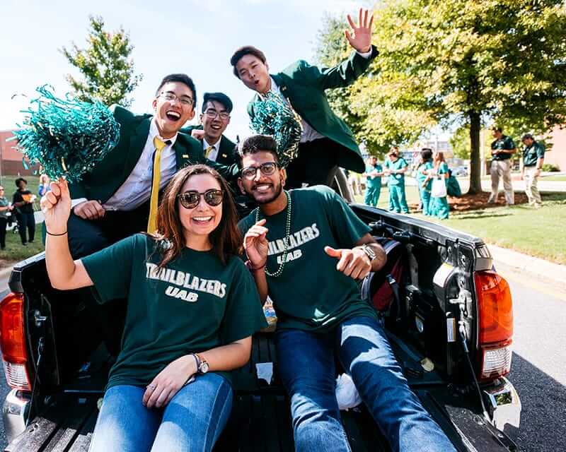 Students riding in the back of a truck at homecoming, smiling and waving at the camera.