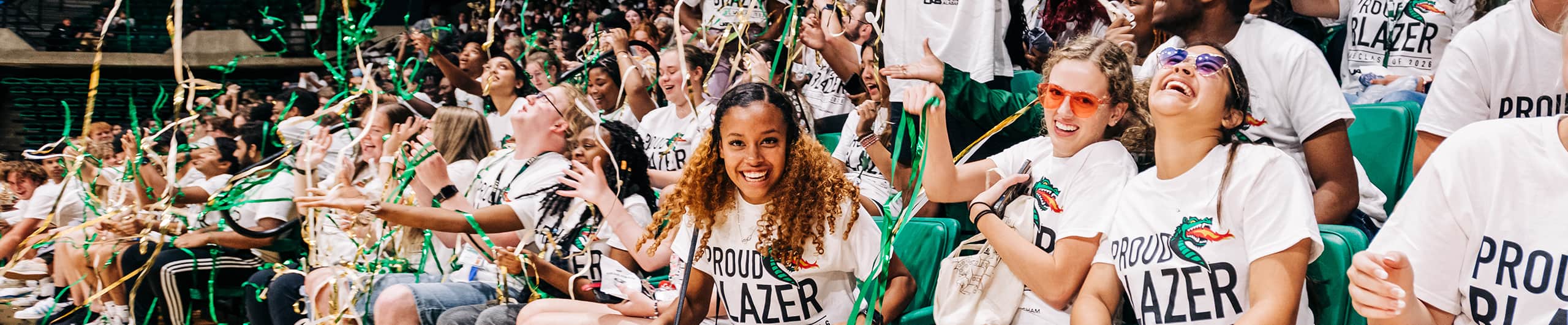 Smiling students at a spirit event wearing "Proud Blazer" T-shirts and throwing metallic green streamers. 