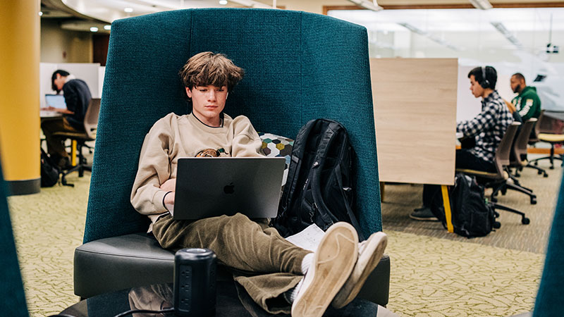 A young man sits in a comfortable chair working on his laptop, his legs propped up on a table. Students in background work at library carousels.. 