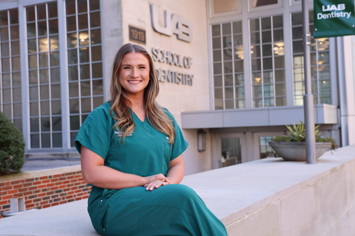 Baleigh Robertson sitting on concrete wall with hands on lap in front of the UAB School of Dentistry Building