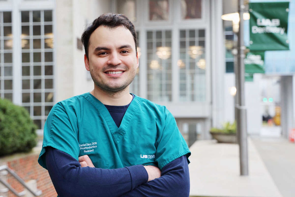 Man in green scrubs and navy blue long sleeves smiling with arms crossed in front of the UAB School of Dentistry