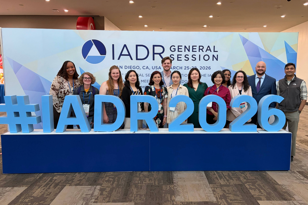 Image of group of people standing behind a large sign that reads, " #IADR2026"