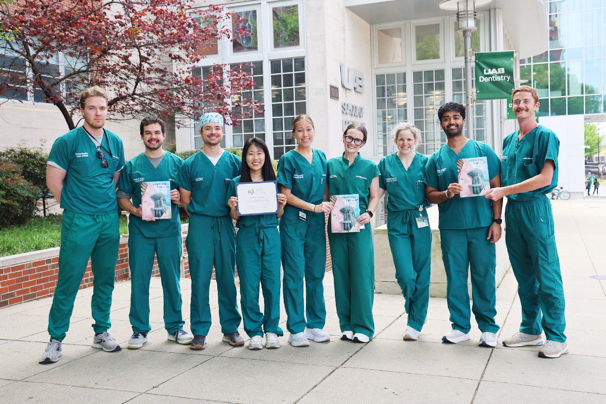 Group of dental students in green scrubs holding their SRG newsletter and certificate in front of the UAB school of Dentistry building
