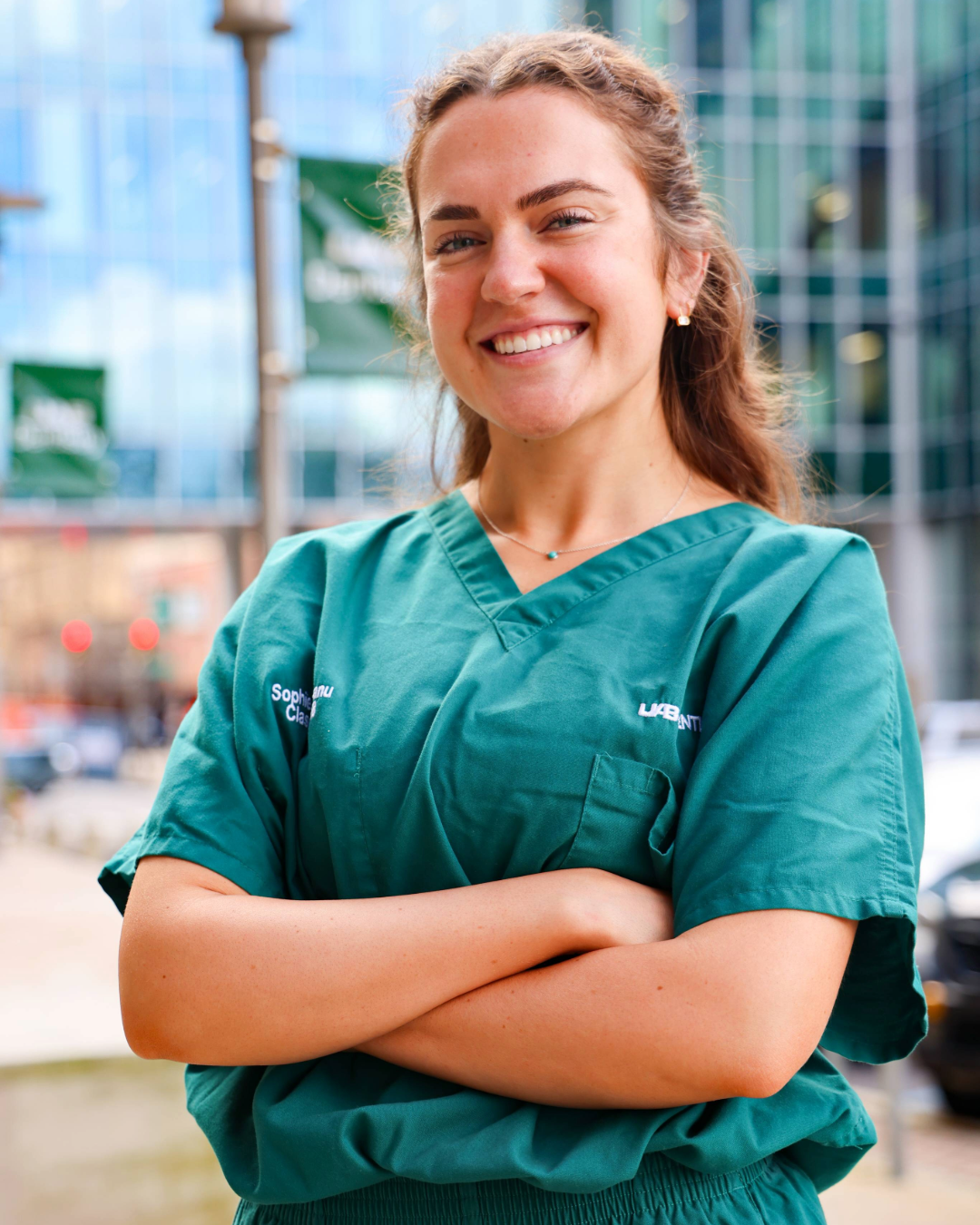 Girl with curly brown hair in green scrubs smiling with arms crossed for a photo in front of a modern metropolitan city setting with glass building and street view. Sidewalk has light poles with green flags.