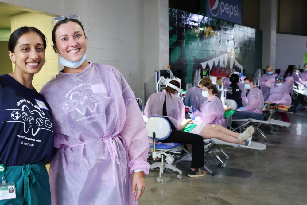 Two students smiling in front of row of students residents and faculty providing dental care at TeamSmile event at Bartow arena
