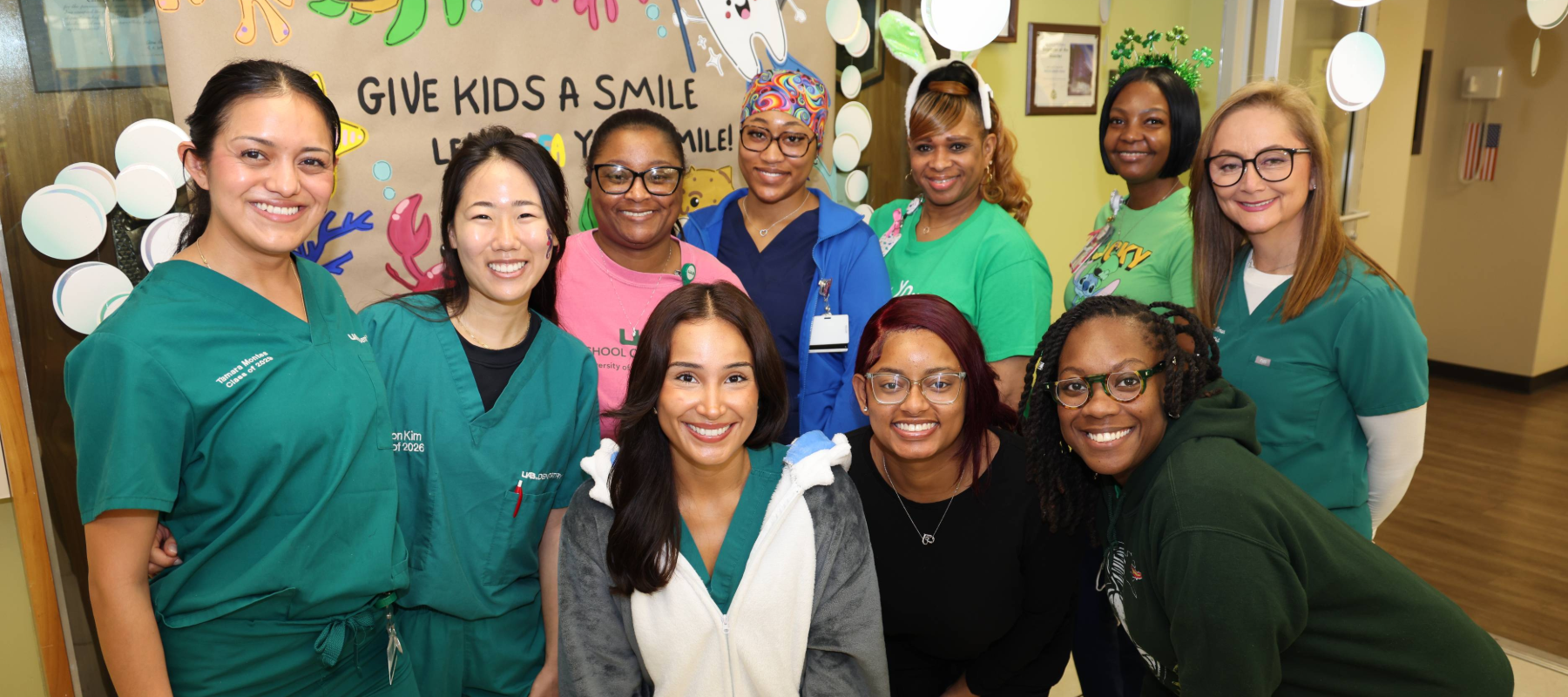 Group photo of dental student, faculty and staff in front of a "Give Kids a Smile" poster in clinical setting