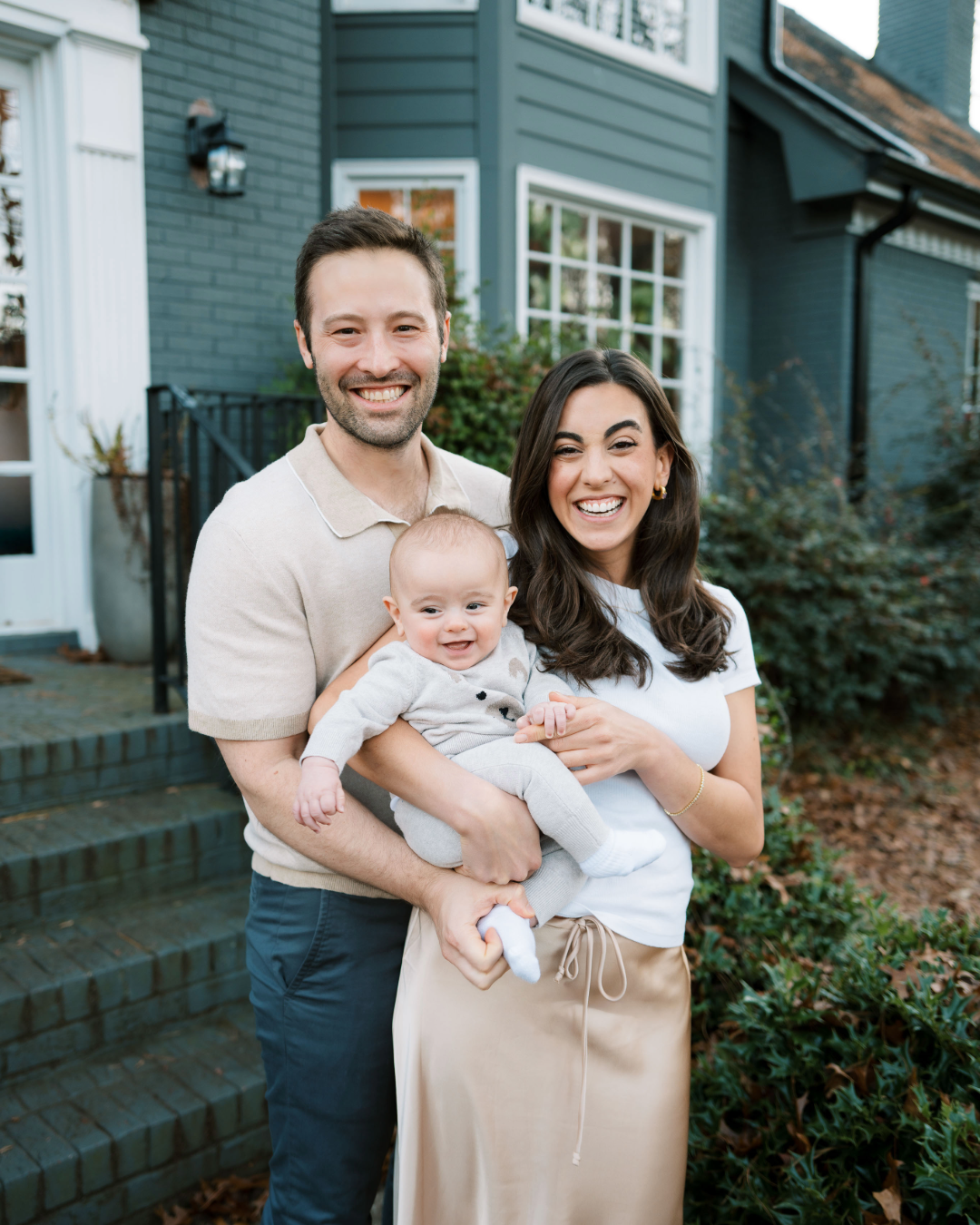 A mother and father holding baby smile for photo in front of modern home
