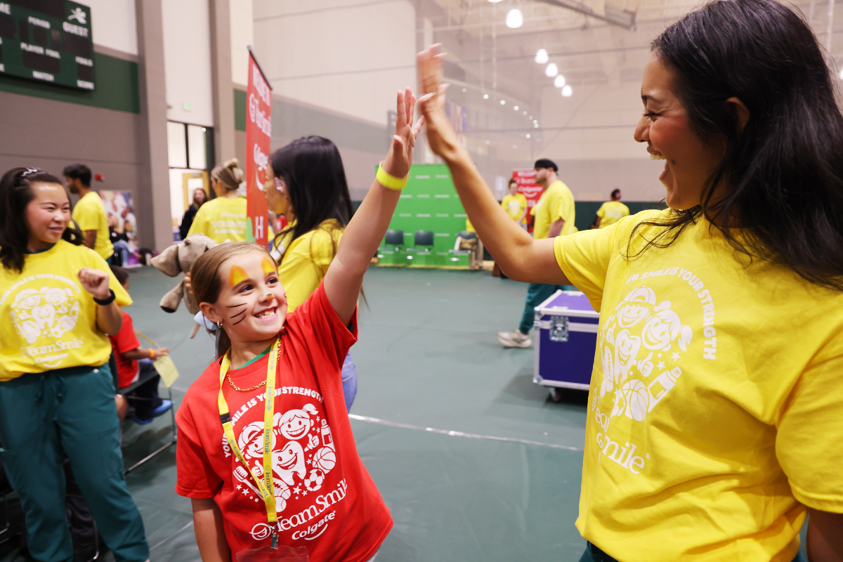 A smiling girl with face-painted whiskers in a red TeamSmile t-shirt high-fives a dental student volunteer wearing a yellow TeamSmile shirt at a lively community event in a gymnasium.