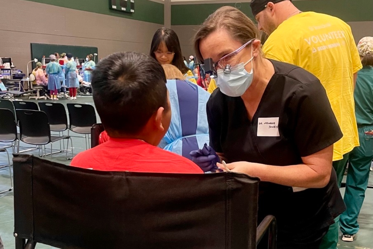 Woman in black scrubs wearing dental loupes and a blue mask consulting with a child during community dental outreach event in gymnasium.