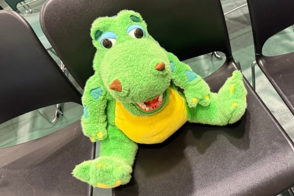 Green dragon plush toy sitting in black chair in gymnasium