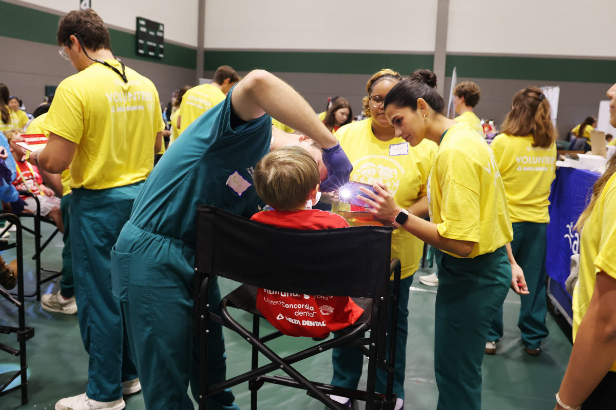 Student dental team and volunteers conducting a community dental checkup for a young child seated in a portable chair, a dental student inspecting the child's mouth with a bright light while other volunteers in yellow shirts assist.