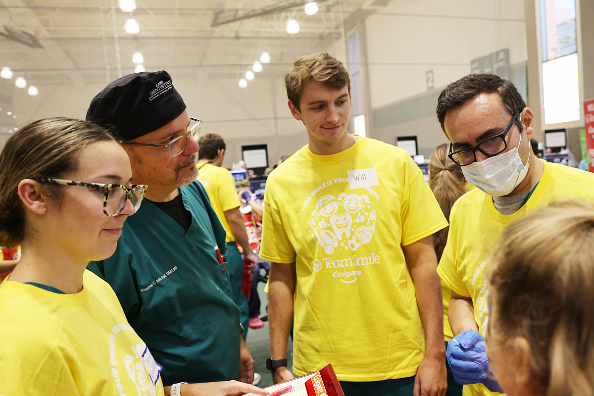 Dental students in bright yellow "Team Smile" shirts and a faculty member, leaning in to speak with a child at a busy community dental clinic.
