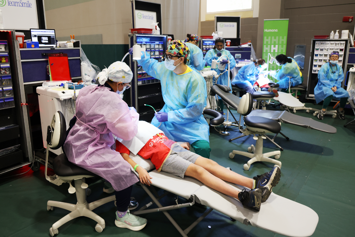Volunteer dental team in PPE providing free dental care to a child reclined on a portable clinic chair inside a community outreach setup with multiple treatment stations.