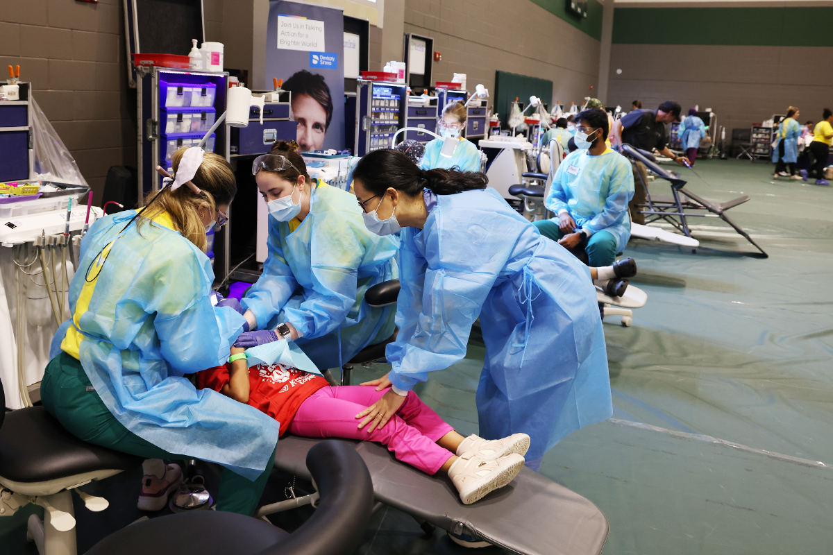 Dental team in blue protective gowns and masks providing dental care to a child reclining on a portable chair during a community dental clinic set up in a gymnasium.