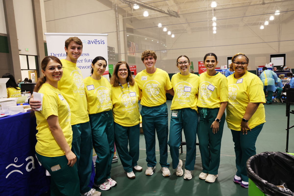 Volunteers at a community dental outreach event stand arm-in-arm smiling, wearing yellow "TeamSmile" T-shirts and green scrubs inside a gymnasium.