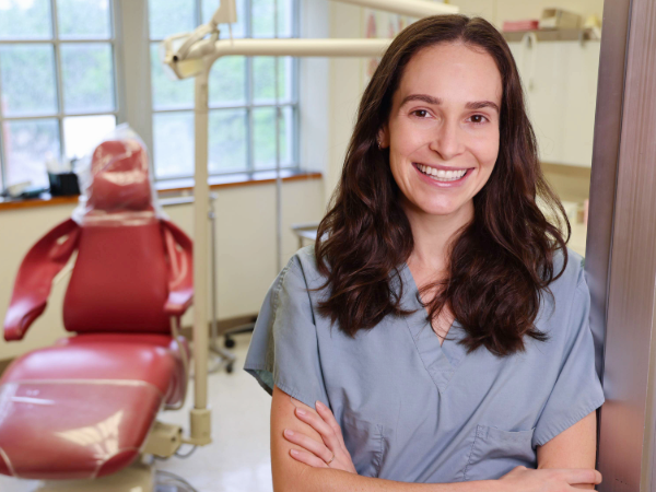 Smiling woman in clue scrubs and arms crossed standing in door way of dental clinic operatory with red chair