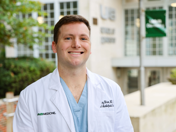 Smiling man in white coat standing outside in front of the UAB School of Dentistry building