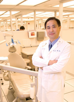 Dr. Liu standing in front of a dentistry chair in the school, arms crossed, wearing a white coat. 