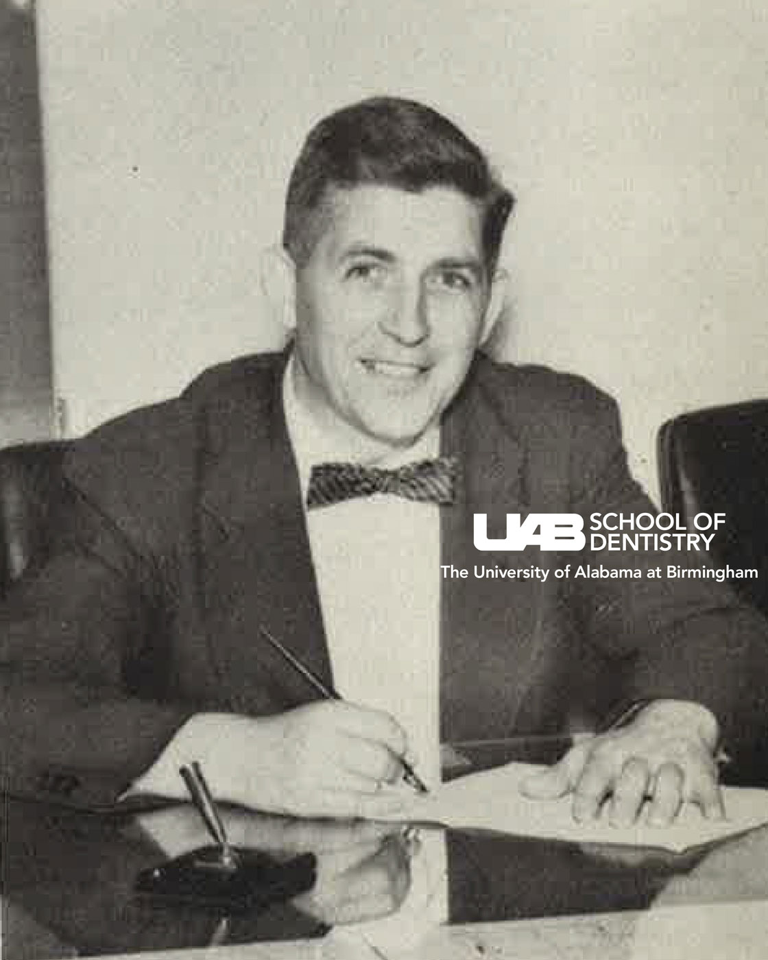 Black and white image of Dr. Volker, wearing a dark suit and striped bow tie, setting a polished desk signing a paper.