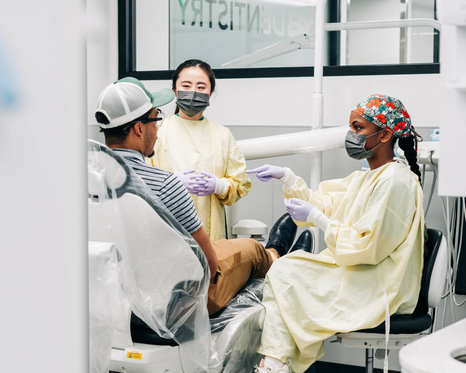 Girl in clinical gown, mask, and scrub cap pointing towards man in dental chair wearing a hat and striped shirt while another girl with a clinical gown and masked looks at him