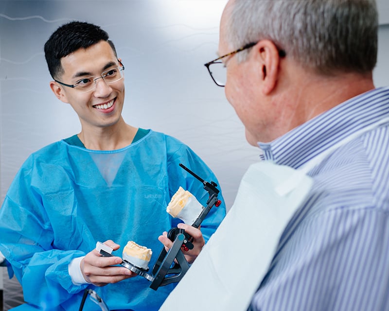 Dentist in scrubs holding a mouth mold and talking to a patient wearing a dental bib.