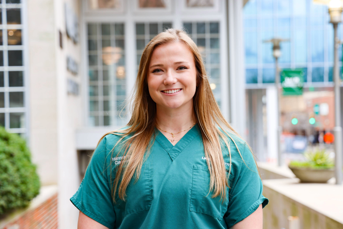 Blonde woman in green scrubs smiles in front of UAB School of Dentistry building.