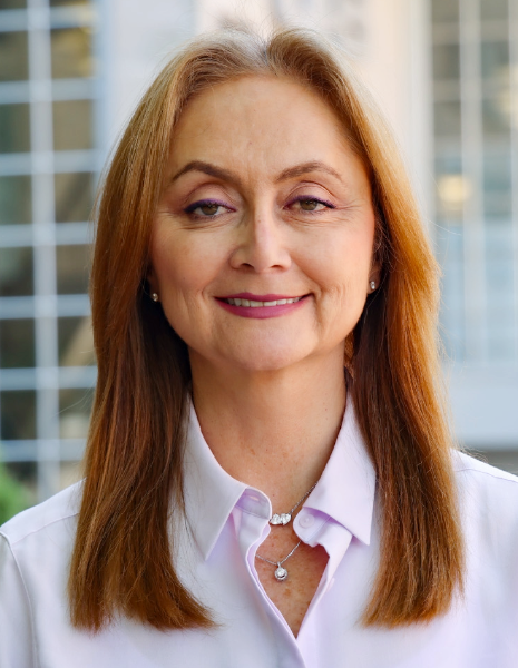 Woman with brown hair and white blouse and diamond necklace smiling for photo in front of building exterior with windows