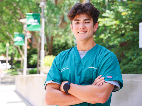Male dental student in green scrubs smiling with arms crossed outdoors