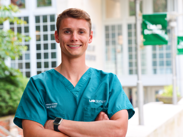 Young smiling dental student in teal scrubs embroidered "UAB Dentistry," standing outside a campus building with arms crossed.