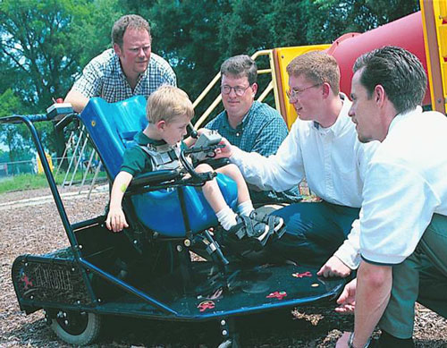 Engineering students talk to a handicapped boy driving his wheelchair with a joystick. 