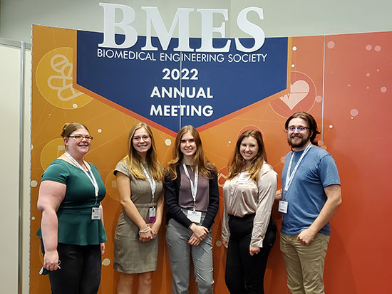 5 students in front of an orange annual meeting screen.