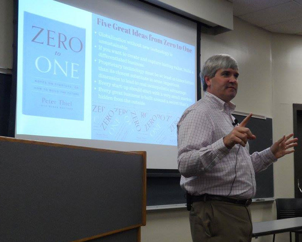 A UAB IEM speaker lecturing at the front of a classroom in front of a screen displaying a PowerPoint presentation.