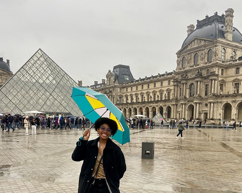 African American female student with a big smile and colorful umbrella stands outside the Louvre. 