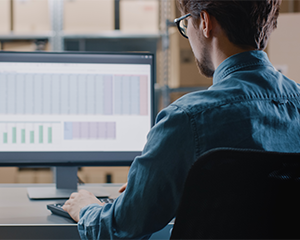 Man sits in front of a desktop computer which displays a spreadsheet