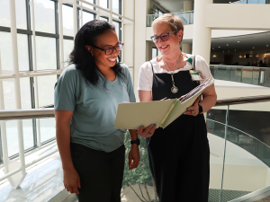 Two women stand in a building at UAB, looking at a file and smiling together.