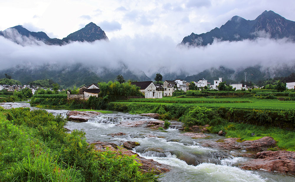 Village nestled below mountains under a low layer of clouds, green grass and a rocky stream in the foreground.